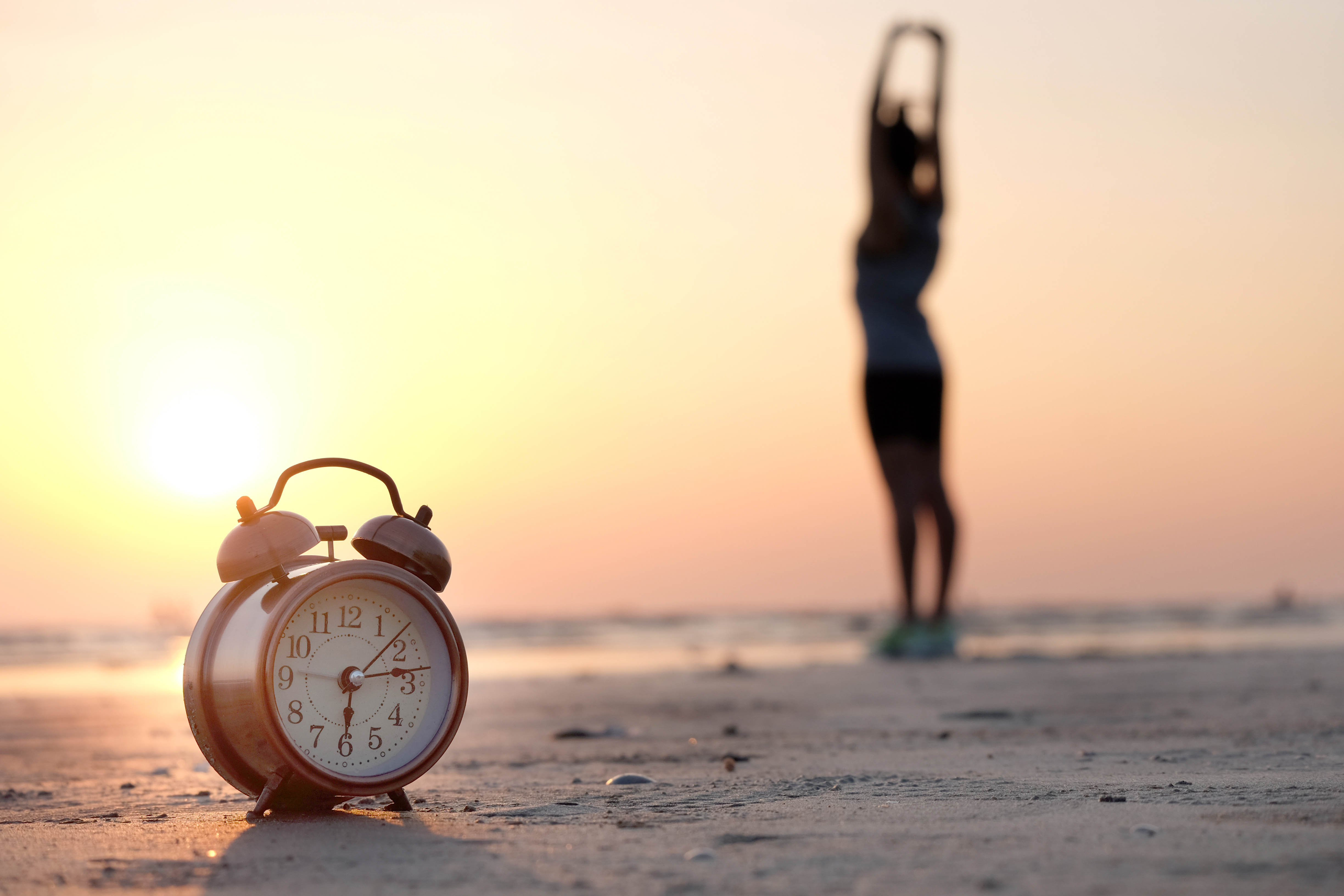 Woman stretching on the beach facing the sunrise, clock in the foreground showing that it is 6:15am Woman stretching on the beach facing the sunrise, clock in the foreground showing that it is 6:15am